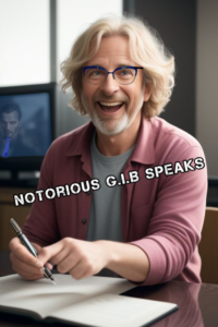 A smiling middle aged man wearing blue rimmed glasses, moderate wavy grey hair (collar length) holding a pen while sitting at a desk. He is wearing a light blue shirt with a pink coloured shirt over top. NOTORIOUS GIB SPEAKS is stamped across the middle of the picture.