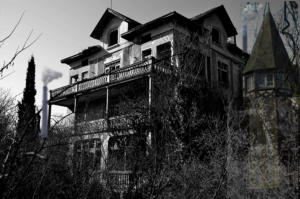 A black and white picture An old 4 story house. The shot is taken from off to the right side of the house. There is overgrown shrubs and trees.  Some windows appear to be without glass. There are a couple of smoke stacks from a factory in the background.