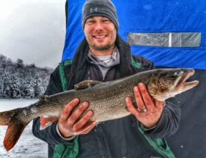 Jeremy smiling while standing on ice with his blue portable ice hut in the background holding a good sized laker. He is all bundled out with a touque and winter jacket. His fingers are slightly red.