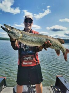 A beautiful sunny day. Jeremy is standing in his boat, wearing his dark sunglasses, ball cap, and sponsors shirt. He is holding a "trophy sized" musky almost at shoulder height.