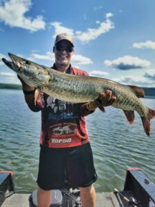 A nice sunny day. Jeremy standing in his boat holding another monster musky wearing his ball cap, dark sunglasses, and sponsor shirt.