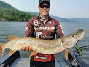 Jeremy standing in his boat holding a monster musky at waist height. He is wearing his dark sunglasses, ball cap and sponsors shirt. A green and silver musky lure is still hanging from the mouth of the fish.