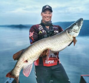 Jeremy standing in his boat. A nice day and the water very calm.. almost like glass calm. He is wearing a dark ball cap, and his sponsors shirt while holding a huge musky slightly on an angle.