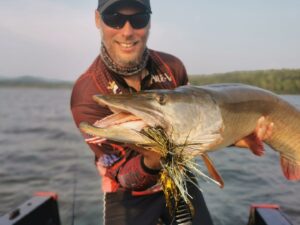 Jeremy in his boat, wearing dark sunglasses, his ball cap and sponsors shirt while showing the open mouth of a musky