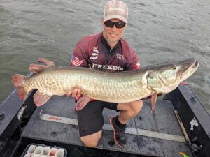 Jeremy, wearing dark sunglasses, a ball cap and shirt with his sponsors listed on it holding a good sized musky in his boat.