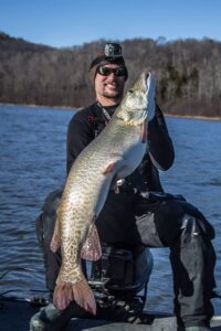 Jeremy sitting on the outboard motor of his boat wearing a touque with his Go-Pro camera attached, sporting dark sunglasses and holding vertically his first 50" musky.