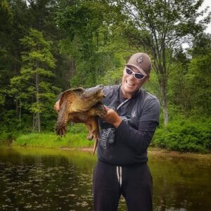 Jeremy in the water holding a large snapping turtle by the sides of it's shell. Jeremy is laughing, wearing his white rimmed dark sunglasses, ball cap, and sponsors shirt. The turtle has it's mouth wide open and does not look impressed.