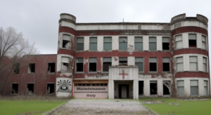 An abandoned building. Three floors and a garage door to the left of what looks to be the main entrance. Building looks to be either that of a school or an old hospital. There is a medical cross over the front door. 