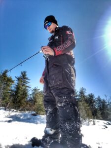 Jeremy, dressed for warmth with a touque and sunglasses. Hes standing on the ice, not far from shore on a bright sunny winter day folding his ice fishing rod.