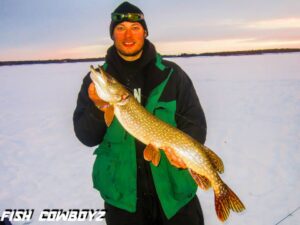 Jeremy standing on a frozen lake, dressed warm and holding a good sized pike that he just caught.