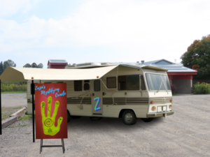 A off white coloured Winnebago with brown stripe down the side parked in a dirt parking lot. The Winnebago has a Z on the door and the awning out and extended with a sign out in front advertising Zaza Mystifying Oracle.