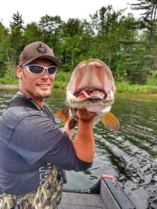 Jeremy wearing a ball cap and sunglasses on his boat holding up a musky. The musky is facing the camera with it's mouth wide open.